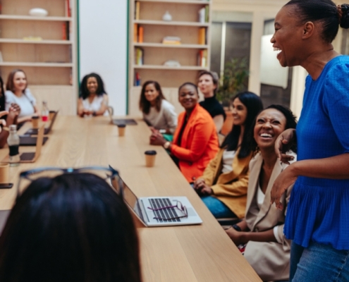 A large group of people sit around a conference table in a casual setting. A 30yo black woman is standing, in the foreground, speaking and laughing. The attendees are engaged and listening, smiling