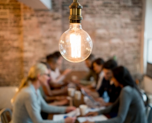 soft focus image of a diverse group of people sitting at a table, talking. Papers, laptops, and cups of coffee are in front of them. A modern light fixture featuring an Edison-style bulb is centered and in focus, with the working group behind/below.